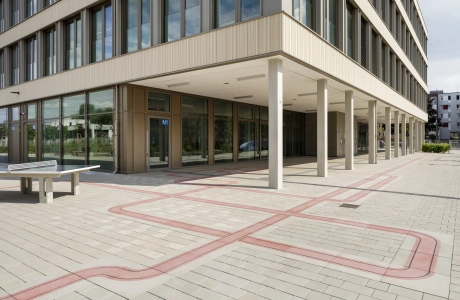 Ein Schulhof, welcher zum Teil vom großen Schulgebäude überdacht wird. Gepflastert ist er zum Teil mit grau-beigen, versickerungsfähigen Pflastersteinen und zum anderen mit roten Betonsteinen, welche sich wie ein Kunstwerk durch die Fläche ziehen.