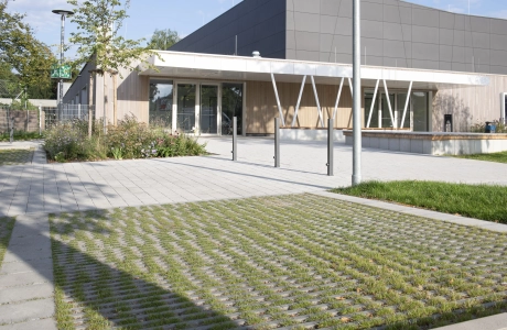 Im Vordergrund ist ein Parkplatz aus begrünten Rasenfugensteinen. Dahinter ist ein Weg – gepflastert mit dem versickerungsfähigem Klimastein – und eine moderne Sporthalle mit anthrazitfarbener Fassade und Holzverkleidung. Drumherum sind grüne Bäume und Sträucher zu finden.
