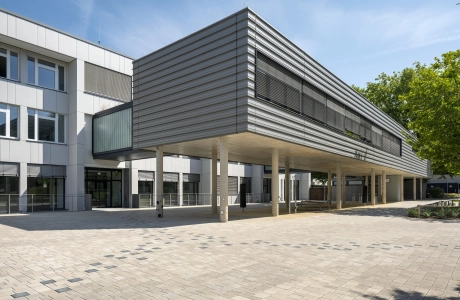 Open schoolyard, which is rounded off on the right-hand side with a meadow and trees. A path consisting of anthracite-colored concrete blocks runs through the paved courtyard to a modern, multi-part school building. The front part stands on concrete columns and is clad in dark gray. The rear part has a white façade. 