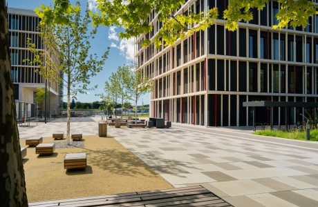 Moderner Campusinnenhof mit gepflastertem Platz, jungen Bäumen und Sitzbänken, umgeben von mehrstöckigen Bürogebäuden mit großen Fensterflächen bei sonnigem Wetter.