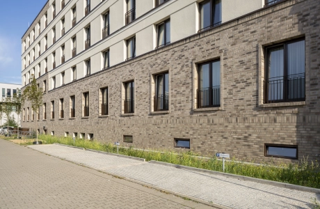 Moderne Gebäudefassade mit beigem Anstrich im oberen Bereich und braunen Wandfiesen im unteren Bereich. Davor ist ein gepflasterter Parkplatz und ein Weg. Zwischen der Parkfläche und dem Gebäude ist ein Grünstreifen zu finden.