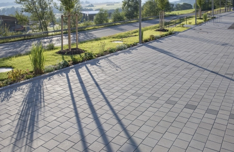 Nahaufnahme von grauem, versickerungsfähigem Betonsteinpflaster. Im Hintergrund ist eine hügelige Landschaft mit Sträuchern, Gräsern und Bäumen zu erkennen.