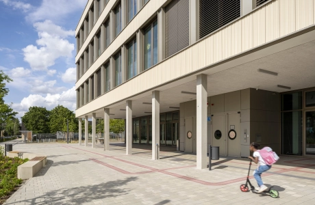 Fassade eines Schulgebäudes mit großen Fenstern und Säulen. Davor liegt ein grau-beigen geplasterter Platz mit Sitzelementen und einem Kind, welches mit einem Roller fährt. Im Hintergrund sind viele Bäumen zu erkennen.