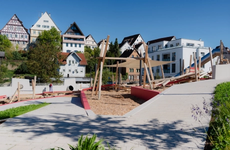 Moderner Spielplatz aus Holz mit Klettergerüsten, Schaukel und Rutschen, umgeben von gepflasterten Wegen und Grünflächen. Im Hintergrund ist eine Altstadt mit alten Fachwerkshäusern zu sehen.