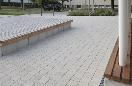 Nahaufnahme von Kalk-Grauen, versickerungsfähigen Betonsteinen. Daneben befindet sich eine Sitzbank aus Beton mit Holzlatten als Sitzfläche. In dieser enden mehrere weiße Stützen. Auf der anderen Seite befindet sich ebenfalls eine Sitzmöglichkeit aus Beton mit hölzerner Sitzfläche.