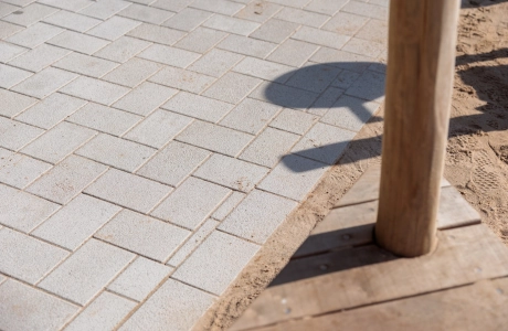 Nahaufnahme eines hellen, beigefarbenen Pflasters aus GDM.KLIMASTEIN. Rechts im Bild befinden sich ein Holzpfosten und ein hölzerner Randabschluss, daneben liegt ein angrenzender sandiger Bodenbereich.
