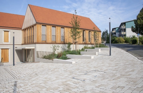 Modernes Gebäude mit Holzfassade und Ziegeldach neben einem ökologischem Flächenbelag mit Bänken.