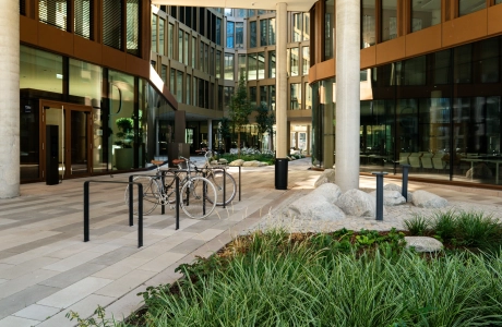 Blick auf den Innenhof eines großen Gebäudekomplexes mit vielen Fenstern. Der Innenhof ist mit hellem Betonstein gepflastert. Im Vordergrund ist ein grün bepflanztes Beet und daneben befinden sich Fahrradständer mit abgestellten Fahrrädern.