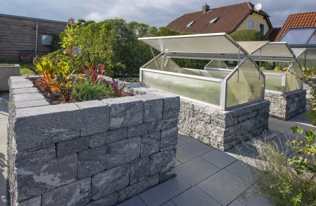 3 klassische Hochbeete aus grau-schwarzen Betonsteinen. Im vordersten Beete wächst grün-roter Salat. Die beiden anderen Hochbeete haben ein integriertes Gewächshaus. Im Hintergrund sind Bäume und Häuser zu erkennen.