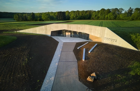 Modernes Gebäude mit Holzoptikfassade und großer Fensterfront. Davor ist ein Weg, welcher auf das Gebäude hinzuführt. Drumherum sind erdige Flächen, auf denen Betonelemente zu finden sind.
