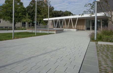 Weg aus Kalk-Grauen, versickerungsfähigen Klimasteinen, welcher zu einer Sporthalle hinführt. Diese hat eine weiße Überdachung, welche durch schräg aufeinander zulaufende Träger gehalten wird. Drumherum finden sich Grünflächen mit Sträuchern und Bäumen.