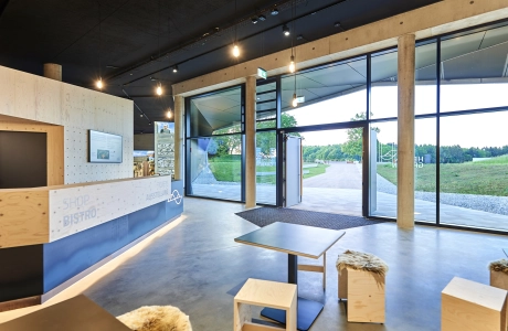 Modern gestaltetes Foyer mit großer Fensterfront. Der Empfang und die Sitzmöglichkeiten sind aus hellem Holz und anthrazitfarbenen Designelemente