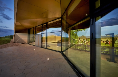 Große Fensterfront eines modernen Gebäudes. Davor sind fünfeckige, kalksteinfarbene Betonsteine verlegt. 