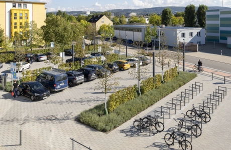 Blick von oben auf Parkplätze mit Autos und Fahrradstellplätze mit Fahrrädern. Zwischen den Parkreihen sind Beete mit grünen Sträuchern, Gräsern und Bäumen. Im Hintergrund sind mehrere Gebäude zu erkennen.