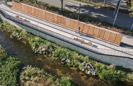 Luftaufnahme eines Uferwegs entlang eines flachen Bachlaufs, gesäumt von Holzsitzbänken und einer langen Holzverkleidung. Der Weg ist mit hellem Betonstein gepflastert, während oberhalb eine Straße mit Bäumen verläuft.