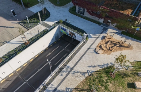 Luftaufnahme eines modernen Außenbereichs mit einem gepflasterten Weg aus hellem Betonstein, angrenzend an einen Straßentunnel, ein begrüntes Gebäude und einen Sandspielplatz.