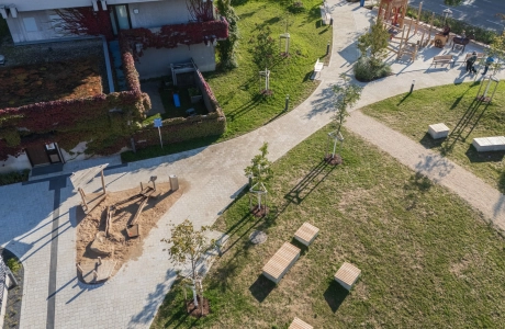 Luftaufnahme eines neu gestalteten Außenbereichs mit geschwungenen Wegen und großzügigen Grünflächen. Der zentrale Weg sowie der Vorplatz bestehen aus beigem GDM.KLIMASTEIN. Sichtbar sind mehrere junge Bäume, Sitzbänke aus Holz, ein kleiner Sandspielbereich mit Holzspielgeräten und ein weiterer Spielbereich mit Kletterelementen auf der rechten Seite. Im Hintergrund stehen begrünte Gebäude und entlang der Wege sind einzelne Personen erkennbar.