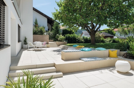 Terrassenplatten in einem begrünten Garten mit einem großen Baum und Sitzelementen aus Beton.