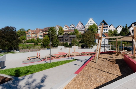 Moderner Spielplatz aus Holz mit Klettergerüsten, Schaukel und Rutschen, umgeben von gepflasterten Wegen und Grünflächen. Im Hintergrund ist eine Altstadt mit alten Fachwerkshäusern zu sehen.