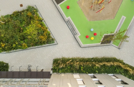 Draufsicht auf eine mit basalt- und granitfarbenem GDM.SCENIC individual gepflasterte Fläche. Zwischendrin sind begrünte Beete und am Rand ist ein großes Wohngebäude zu finden. Außerdem gibt es eine erhöhte Fläche mit grünem Bodenbelag und einem bunten Spielplatz.