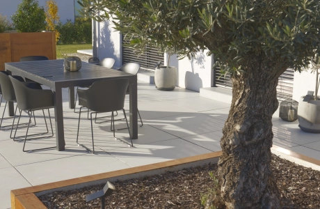 Terrasse auf der GDM.MASSIMO light verlegt ist, mit einem großen Baum mit Cortenstahl‑Beeteinfassung und einem dunkelgrauen Outdoor-Esstisch mit Stühlen. Im Hintergrund stehen Pflanzkübel an der Hausfassade.