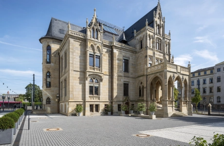 Historische Villa mit mehreren Stockwerken und aufwendigen architektonischen Details. Das Gebäude hat eine helle Sandstein-Fassade, verzierte Fensterrahmen und einen ausgeprägten Eingangsbereich mit darüberliegendem Balkon. Der gepflasterte Platz um die Villa ist gepflegt und von Bäumen und Pflanzgefäßen umgeben. Im Vordergrund sind auch Parkplätze zu erkennen