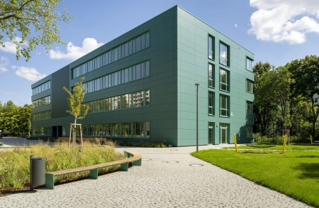Ein mit rundlich gepflasterteten Betonsteinen gepflasterter Weg, welcher direkt zu einem modernen Schulgebäude mit petrolfarbener Fassade führt. Der Bereich links und rechts neben dem Pfad ist mit Streuchern, Wiesen und Bäumen begrünt.