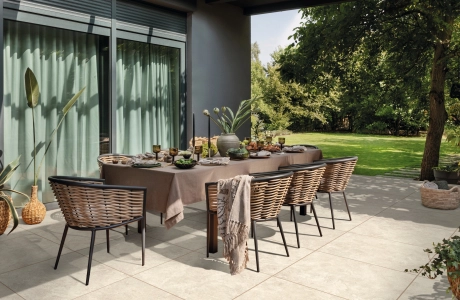 Moderne Terrasse mit schön gedecktem und dekorierten Tisch mit vielen Stühlen und Keramik-Verbundplatten am Boden verlegt. Im Hintergrund ist ein grüner Garten und das Wohnhaus mit großen Fenstern und anthrazit-farbener Fassade zu erkennen.