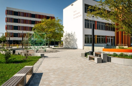 Ansprechender Schulhof mit vielen Sitzelementen und Grünflächen an einem sonnigen Tag mit blauem Himmel. Ein Klettergerüst innerhalb einer Sandfläche ziehrt den Hof und große Bäume spenden Schatten. Im Hintergrund stehen zwei Schulgebäude mit weißen Fassaden und orange-gelben, geometrischen Verzierungen