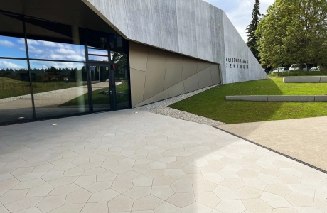 Im Vordergrund sind fünfeckige, kalksteinfarbene Betonsteine verlegt. Dahinter ist eine moderne Gebäudefassade in Holzoptik mit Fensterfront. Daneben ist eine grüne Rasenfläche.