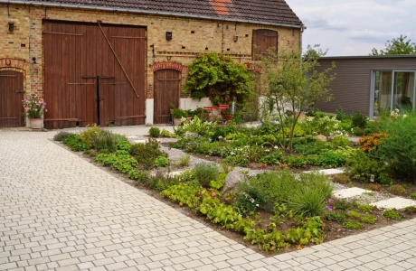 A rustic brick building with large wooden gates and a landscaped garden in the foreground. A paved path leads through the garden, which is planted with flowers and shrubs. In front of the garden is a natural beige paved courtyard laid out in rows. A modern building with large glass doors can be seen on the right.