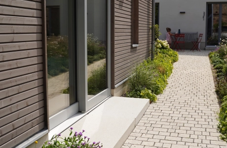 Ein modernes Gebäude mit einer Fassade aus dunkelbraunen Holzpaneelen. Ein Podest ziert den Bereich vor der Eingangstür und ein naturbeige gepflasterter Weg im Reihenverband trennt das Gebäude von einem Garten . Im Hintergrund ist ein Haus mit weißer Fassade und rotem Ziegeldach zu erkennen. 