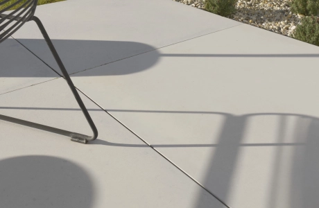 Nahaufnahme heller GDM.MASSIMO light Terrassenplatten, auf denen die Schatten eines filigranen Outdoor‑Stuhls sichtbar sind. Im Hintergrund liegen ein Kiesstreifen, Ziergräser und eine Grünfläche mit einer Steinmauer.