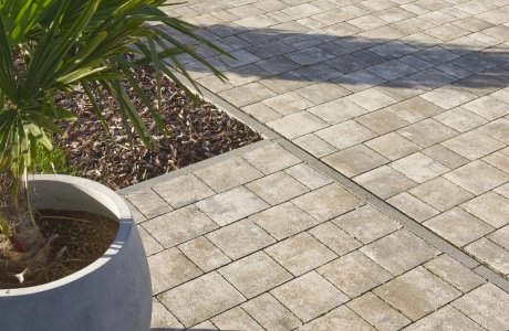 Nahaufnahme des muschelkalkfarbenem GDM.VIA stone. Auf der gepflasterten Fläche steht ein großer runder Pflanztopf mit einer Palmenpflanze, daneben sind Schattenwürfe, eine Beetfläche mit Rindenmulch und Teile der angrenzenden Einfahrt sichtbar.