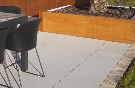 Terrassenbereich auf der GDM.MASSIMO light verlegt ist. Im Vordergrund steht ein Hochbeet mit Cortenstahl‑Einfassung und einem Olivenbaum, daneben ein Outdoor‑Esstisch mit schwarzen Stühlen. Im Hintergrund sind Rasenfläche, herbstliche Bepflanzung und Landschaft zu erkennen.