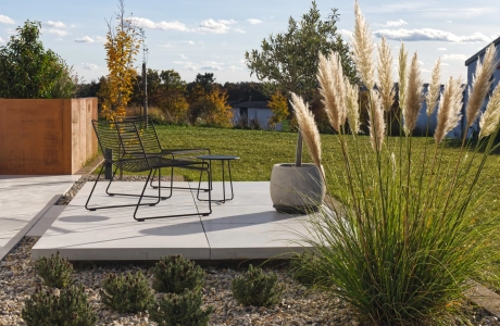 Garten- und Terrassenbereich mit hellen GDM.MASSIMO light Platten, die eine moderne Sitzfläche mit filigranen Metallstühlen bilden. Im Hintergrund umrahmt eine GDM.MASSIMO wall in Cortenstahloptik ein erhöhtes Beet. Davor liegen ein Kiesbeet mit Ziergräsern und eine weitläufige Rasenfläche, eingerahmt von herbstlicher Bepflanzung.