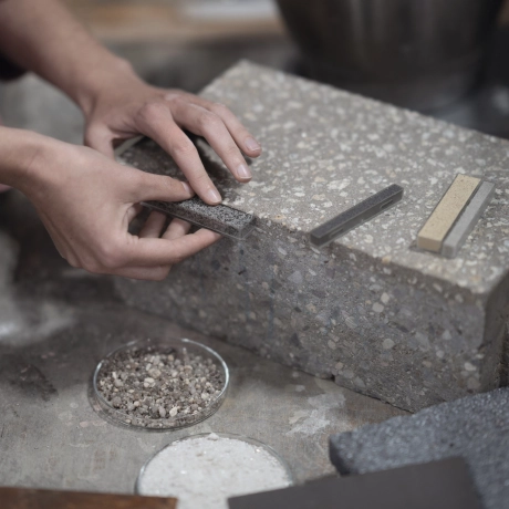 A person compares various material samples with a beige-gray paving stone with light granulation. On the table are a bowl with small stones, another tool, and various materials.
