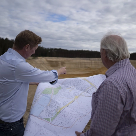 Bernhard Godelmann und Bernhard Godelmann Senior stehen am Rand der Sandgrube mit einem Plan in der Hand und gucken gemeinsam in die Ferne.