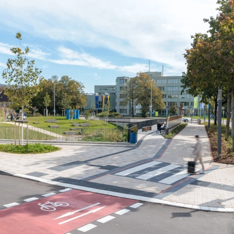 Übersicht einer städtischen Weganlage, in der ein breiter Gehweg überwiegend mit beigem GDM.KLIMASTEIN gepflastert ist. Zur Markierung des Querungsbereichs sind rote und weiße Pflastersteine aus GDM.TETRAGO stone integriert. Die Wege grenzen an eine begrünte Parkfläche mit Rasen, Bäumen, Sitzbereichen und einem Wasserlauf. Im Vordergrund verläuft ein Radweg.