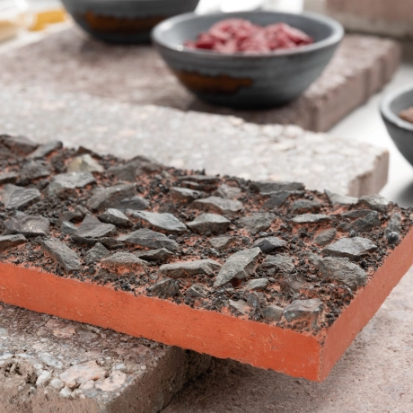 Various handy stone samples for paving stones are visible. In the foreground is a red sample with a rough, textured surface made of black, irregularly shaped granules.