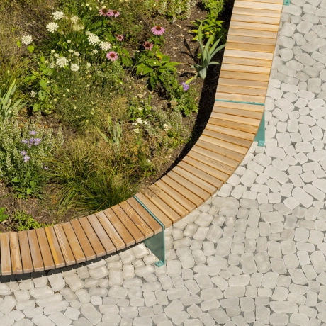 Vogelperspektive auf natürlich und bunt bepflanztes Beet, das von einer Sitzbank mit Holzauflage eingefasst wird. Die Platzfläche ist mit dem rundlich geformten Betonstein GDM.BOLERO stone in zurückgenommenen Grau gestaltet.