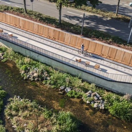 Luftaufnahme eines Uferwegs entlang eines flachen Bachlaufs, gesäumt von Holzsitzbänken und einer langen Holzverkleidung. Der Weg ist mit hellem Betonstein gepflastert, während oberhalb eine Straße mit Bäumen verläuft.