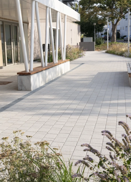 Vorplatz eines modernen Gebäude mit überdachtem Eingangsbereich. Die Überdachung wird durch schräg aufeinander zulaufende, weiße Stützen gehalten, welche in Betonblöcken mit hölzerner Sitzfläche enden. Daneben ist ein Weg, welcher mit Kalk-Grauen, versickerungsfähigen Klimasteinen gepflastert ist. Im Vordergrund sind blühende Sträucher.