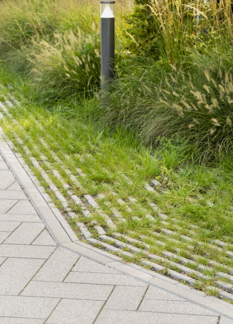 Gepflasterter Gehweg mit basalt- und granitfarbenem GDM.SCADA stone. Am Rand findet sich der begrünte GDM.SCADA liner und verschiedene Gräser und Sträucher. Auch eine schwarze Wegleuchte ist zu sehen.