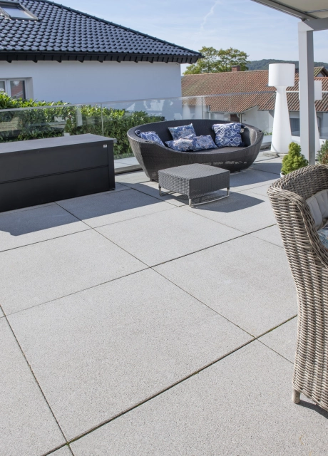 Graue GDM.NUEVA light Platten auf einer Terrasse verlegt. Auf dieser sind verschiedene Gartenmöbel wie eine braune, geflochtene Bank und ein kleiner Tisch.