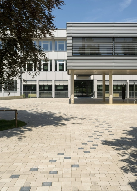 A path consisting of anthracite-colored concrete blocks runs through a paved schoolyard to a modern, multi-part school building. The front part stands on concrete columns and is clad in dark gray. The rear part has a white façade. At the top left, a tree with dark leaves protrudes into the picture.