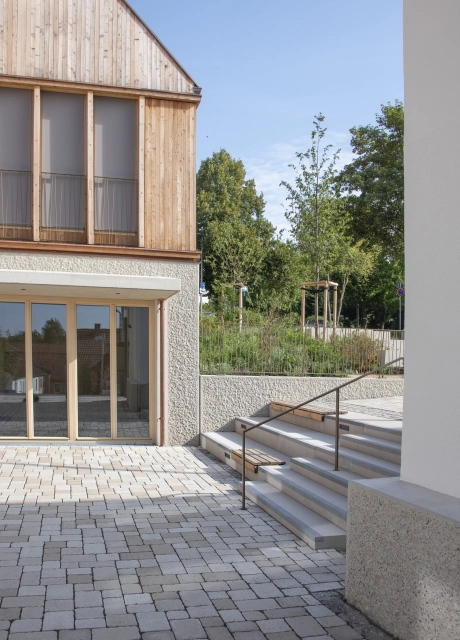 Modernes Gebäude mit oberer Holzfassade, großen Fenstern und ökologisch gepflastertem Innenhof mit Treppe.