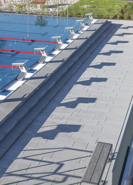 Schwimmbecken mit Startblöcken entlang der Beckenkante und breiten Sitzstufen; auf der umlaufenden Poolterrasse ist der graue GDM.NUEVA stone zu finden.