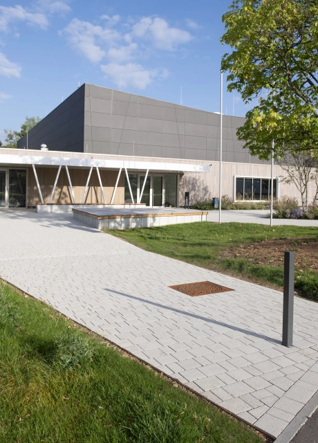 Weg aus dem Kalk-Grauen, versickerungsfähigen Betonstein GDM.KLIMASTEIN, welcher zu einer Sporthalle hinführt. Diese hat eine Fassade mit anthrazitfarbenen Platten im oberen Bereich und eine Holzverkleidung im unteren Bereich. Neben dem Weg befinden sich Grünflächen.