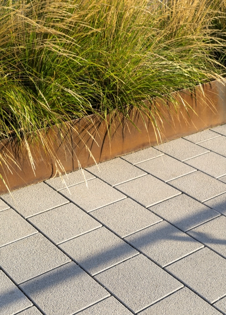 Nahaufnahme der gepflasterten Fläche aus Betonstein. Daneben ist ein Beet mit Gräsern und einer Einfassung aus Cortenstahl.